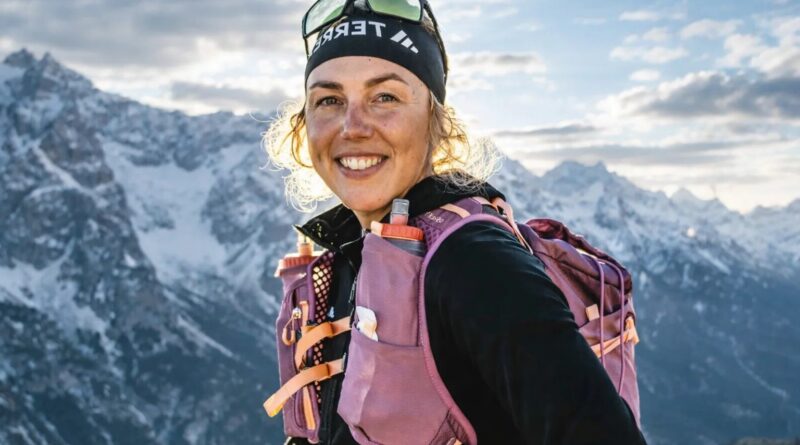 Pirmin Dahlmeier sitzt auf einem Berggipfel in Outdoor-Bekleidung mit Rucksack, im Hintergrund Alpenlandschaft bei Sonnenuntergang, zeigt ein freundliches Lächeln und strahlt Abenteuerlust aus.